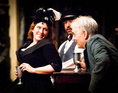 Kate, in velvet-trimmed black dress and black hat with white plume, holds a pewter goblet as she leans back against a table looking over her shoulder at Petruchio, hand on his goblet on the table, leaning forward talking with her; visible between them in the background, sitting at the same table, is Baptista in silver vest, white shirt, and burgundy cravat