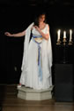 Hermione in Gree-like gown posing as the statue, on a pedestal with a candelabra next to her