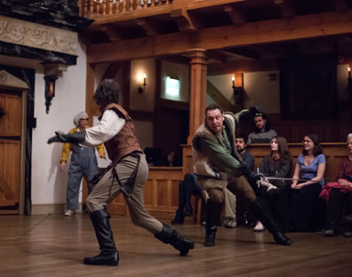 Sword fight with characters, both in vests and knee-high boots; servant in overalls in the corner, and members of the audience behind.