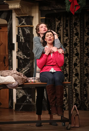 Production photo of Anne Page hugging Courtney Ash, sitting on a table, from behind.