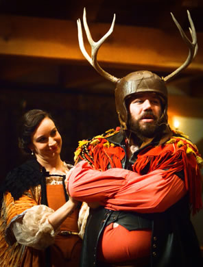 Falstaff wears helmet with a hide covering and 6-point antlers, has his arms crossed waring a tasled red shirt with leath vest, Mistress Ford in elaborat orange dress and white puff sleaves looks sweetly at him