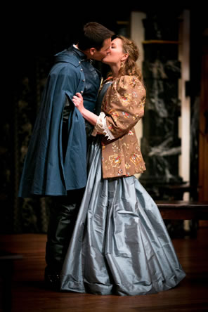 Christian in long blue soldiers coat and black boots kisses Roxane, who is gripping his sleeve, wearing a long blue dress and tapestry-like brown jacket with puff shoulders