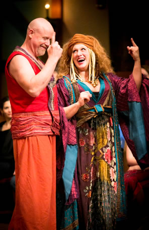 Bolt in orange waste coat and pants (bare-armed) and Bawd in a multi-colored, wildly patterned long silky dress and robe, a brown head wrap and wheat-like cornrows hanging down the side of her head. Both are pointing up with single fingers as they laugh.