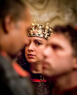 Henry in gold crown and brown leather tunic buttoned at the collar looks on worried at Gloucester and Winchester, nose-to-nose and out of focus in the foreground