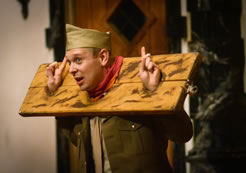 Parolles, in brown army uniform with sky cap, head andhands in stocks while standing up