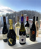 Seven bottles of wine stand on a window sill with windery scene of buildings and a hill of bare trees through the window. 