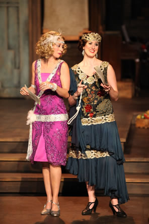 Princess weearing a chartruese dress with sparkly designs, a wide white ribbon with bow as a belt, a sash, pearls and feather tiara holds a letter and looks at the letter Rosalind is holding; she's also holding a string of pearls and is wearing a floral pattern dress with draping gray fabric over the legs and a gold tiara.