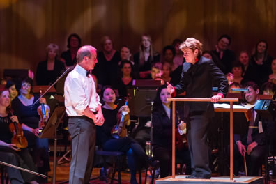 Alsop in a black pants suit stands on the podium, one hand on the railing, as white-shirted Bolger stands meekly, hands at waist, as violinists and choral members watch in the background