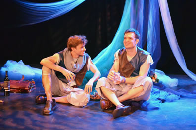 The two cousins sit on the floor, cross legged, in shin-length dingy white pants, brown and blue vests and sandals, Arcite is wearing a gray knit scarf, both holding wine glasses, to one side a leather brief case and wine bottle, in the background the streams of fabric from the maypole