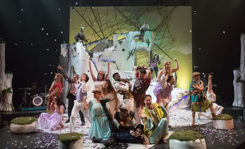 The whole cast in various poses and various costumes, confetti coming down, the stone set in the background in front of a yellow sky and web-like sticks sculpture, the four white boulder around the floor, and two white tree trunks to each side of the stage. 