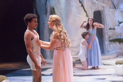 Lysander in white union suit underwear backs away from Hermia wearing a pink gown with her palms on his chest. Int he background, Demetrius, also in white union suit underwear, is on he knees with his hands and face on Helena's waist as she stands in a blue gown with hands up in flustered frustration.