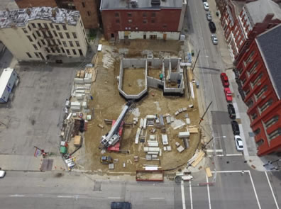Concrete outlines of the thrust stage and rooms to its right amid a dirt covered block with a crane and other construction gear in an overhead photo