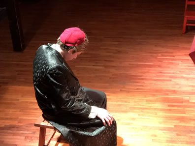 Shylock in black cloak and red skullcap sits on a bench