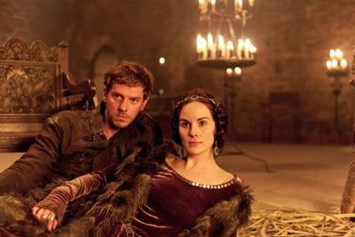 Hotspur in brown leather laies next to Kate in fur cape over borown dress on thrushes in a candlit castle room
