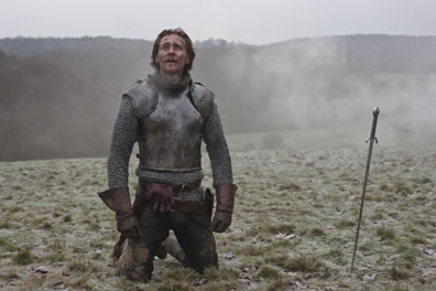 Henry in armor on his knees on the top of a trampled meadow hill with his sword stuck in the ground nearby, and the soldier's gage in his belt