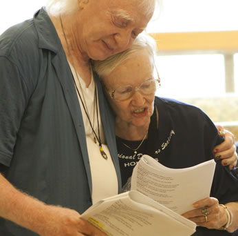 Condos with blue shirt unbuttoned and a white t-shirt underneath, necklaces hanging down around his neck and long white hair hugs Fairchild to his breast  with his left arm around her shoulder, she wearing wireframe glasses and a black t-shirt as she reads a script; both hold scripts in their other hand.