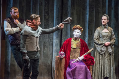 Richad sits on throne, crown o head, scepter in left hand, orb in right, red cloak, purple dress, gold blouse, white ace, Bagot leaning against a wall to the left, arms crossed, Mowbray with glove clenched in outstretched han, silver chainmail like tunik over gray sweater and pants, Elizabeth in patterned silver dress and hands folded in front of her to the right.