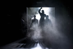 Coming through a large loading dock door with fog rolling in and a bight white spotlight from behind a woman in an overcoat, right fist raised, with other silhouette figures around her