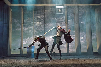 Production photo of Richard, with sword in hand, ducking out of the way of a sword swipe by Richmond, who is wearing an Elizabethan jerkin, skirt, and red cape. 