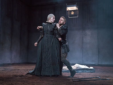 Production photo of Richard in black jacket and pants behind Lady Anne in simple black Elizabehtn dress. His hands are around his shoulder and his leg is crooked, the two crutches on the dirt bloor at his feet. Her head is turned looking at him. In the background is the white shrouded corpse of Henry VI. 