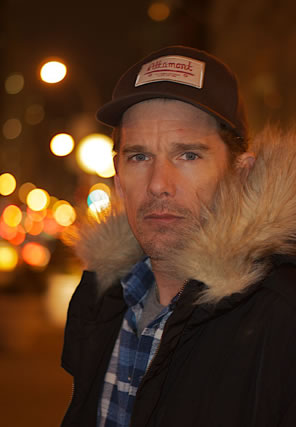 Ethan Hawke in a fur-line hooded jacket and farmer's cap on a city street at night.