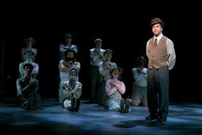 Stage Manager in bowler hat, brown vest and white tie over white shirt and gray pants, behnd him the rest of the ensemble, masked and all with their arms folder over their chests, in three rows: three sitting with legs outstretched in the front, four kneeling, and three standing.