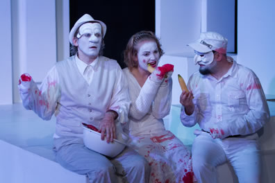 Marcus in fodor hat and vest and white face on the left holds a bowl; Lavinia in the center in white faced gnaws on an unpeeled banana; Titus  in commedia mask and soldiers hat holds a banana. He and Marcus are missing a hand, Lavinia missing both hands, and blood stains are on their white clothes.