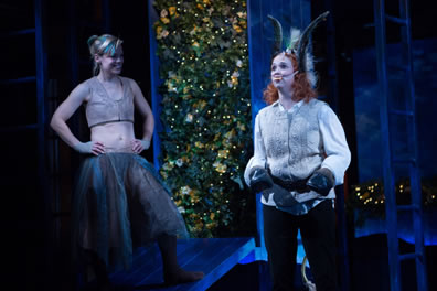 Puck, hands on hips, stands on a platform laughing at Bottom with donkey ears, buck teeth strapped around her cheeks and hooves for hands, wearing a white sweater vest over white shirt with black pans. A bush of vines and fairy lights is in the background, and a ladder to the right