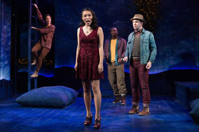 At the front center is Helena in red patterned miniskirt and high heels, at the back left is Hermia climbing down a ladder in brown patterned smock-type shirt and corduroys, in the background are Demetrius in cargo pants, boots, purple jacket and yellow t-shirt, and Lysander in purple leather pants, boots, checkered shirt, denim jacket and fodora. The blue stage has large throw pillows around and fairy lights.