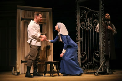 Claudio in white tunic and brown pants and boots holds hands with Isabella in white headscarf and long blue dress, standing up from kneeling, with Duke in green scarf covering his head listning through the gate. The jail door is behind Claudius, and there's a foot stool in front of him.