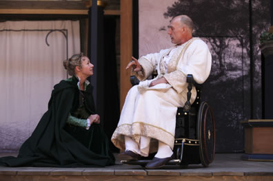Helen in black overcoat kneels to the king dressed in a white robe and sitting in a wheelchair