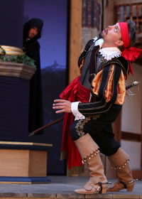 Parolles in cavilier clothes, puffy black and gold sleeves, red sash, tan high-heeled riding boots, red bandanna, reacts to a noise above him while a hooded man peers at him from behind a column in the background.