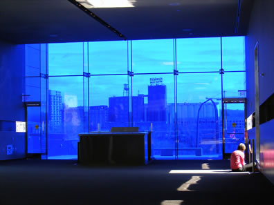 A large plate glass window looking out onto a skyline, lights coming through other windos, one illuminating a woman sitting on the floor writing in a book.