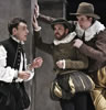 Hamlet in bulging black Elizabethan-style breeches, black vest and high collar white shirt to the left talks with Rosencrantz in green and gold breeches and jacket with white ruff collar dog cap and Guildenstern in gold pants brown jacket and lace collar with country brimmed hat, both leaning against a wall