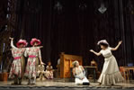 Two guys playing the stepsisters hold up a frame with two flower patterned dresses on them as Cinderella kneels cleaning on the floor and the stepmother in a frilly white dress holds her hands out in incredulity. Piano and other actors in the background