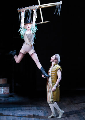 A fairy in vaudeville like lingerie hangs from a chandelier as Puck, in longcoat and britches, talks with her.