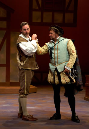 Grumio in brown striped britches and jacket over white shirt, knee socks and brown shoes and a satchel over his shoulder holds the fist of Petruchio in Elizabethan blousy pants of gold and black, with a turquoise waist coat over gold brocade shirt and a purple felt cap, a sword at his side and cape over shoulder