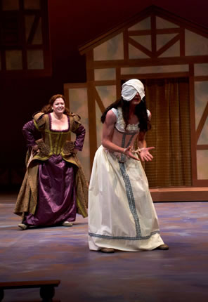 Katherina in purple Elizabethan dress with gold brocade overlay runs toward Bianca in white underdress and tan girdle, her hands bound and head covered in a cloth.