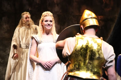 Simonides in background in cream robe and curly beard, Thaisa in white gown and blond tressels, her hands folded at her waist and smiling as she looks on the shield the knight, his back to us, is holding up, he wearing a greenish gold breastplat and simple, gold, conical helmet.