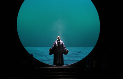 Gower in robes stands with hands outstretched in the middle of a giant circle with a sea behind him and steps leading down to the stage