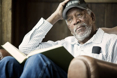 Freeman with white, close-cropped beard, wearing blue-striped white shirt, blue jeans, and a baseball cap that says "One Six Right: The Romancer of Flying", book opened on lap, right elbow resting on chair arm with hand on head, he's looking off in the distance.