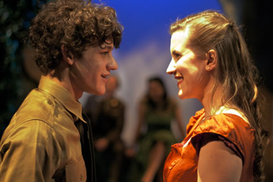 Facing each other, curly-headed, shy Claudio stars at radiantly smiling Hero while other cast members look on in the background