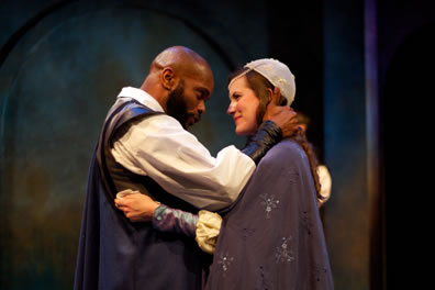 Othello with bald head and beard and blue cape embraces with Dessdemona in cap, blue cape and jewelled gown