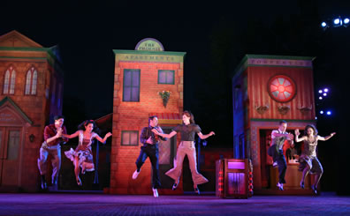 Three pairs of dancers in air as they leap doing the jitterbug, the woman in the center pair wearing slacks. In the background are three of the towers of the set, one a church, one the Phoenix Apartments, and one the Porpentine, and a jukebox sits on the stage, too.
