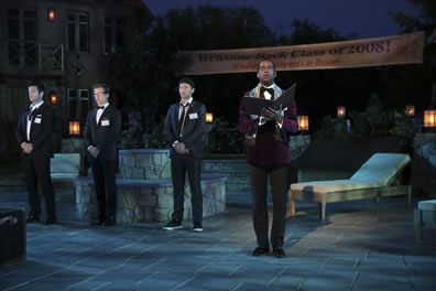 The king in red formal dinner jacket with open book, the lords in tuxedoes (Berowne's bowtie untied,Longaville with backward baseball hat and sneaker) on a flagstone pattio, with lounge chairs, and a banner welcoming class of 2008.