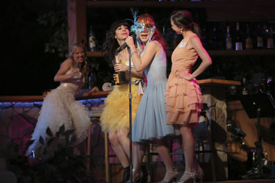 Princess in long white, frothy prom dress sitting at the bar, Rosaline in knee-length blue cocktail dress and mask at a microphone with, on either side of her, Katherine in yellow duck down type dress and holding a bottle of wine, and Maria in short, multi-fold peach dress.