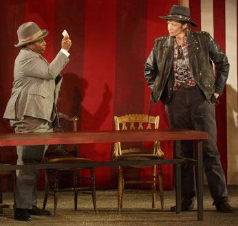 Baptista, wearing a tan suit and fedora, holds up a wad of cash before Petruchio in leather jacket, unbuttoned checkered shirt, death's head t-shirt, black jeans, and black outback hat. They are standing behind a table with chairs. 
