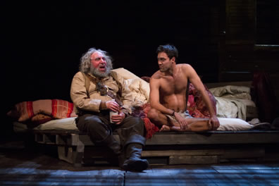Falstaff in gray beard, cream shirt and tan vest with brown pants and black boots holdig a white bottle sits on the edg of a rumpled bed next to Hal, naked except for his briefs