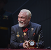 Patrick Page as King Lear, wearing a dark blue suit with purple tie, his jacket adorend with medals and a med on a red and blue ribbon around his neck, sits at a desk, arms folded and sporting a gentle smile. In front of him on the desk is a clock-centered pen stand. Photo by DJ Corey Photography.