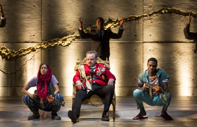 Macbeth slouches in a low-back thrown, wearing a red uniform jacket with medals, fringed shoulder braids and sash. On both sides, two young soldiers squat, both holding AK-47 rifles. the boy in aqua pants, striped shirt and sweater tied around his shoulders, the girl in striped blued pants, purple tea, and red hajib. In the background, Fleance in a suit has his arms raised.